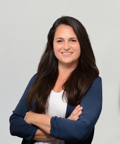 Sandra Unger, ekaflor-Mitarbeiterin Sandra Unger - Frau mit langen braunen Haaren, weißem Oberteil und dunkelblauem Blazer vor weißer Wand